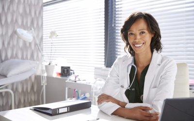 Portrait Of Smiling Female Doctor Wearing White Coat With Stethoscope Sitting Behind Desk In Office